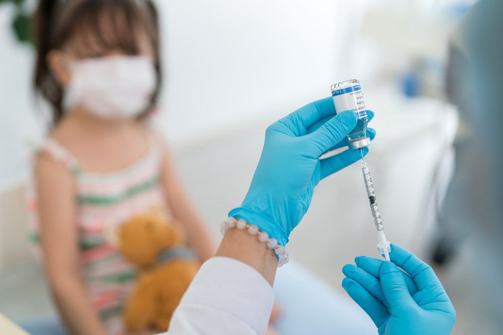 A masked young child with dark brown hair holds a teddy bear in the background while a doctor draws liquid into a syringe in the foreground.