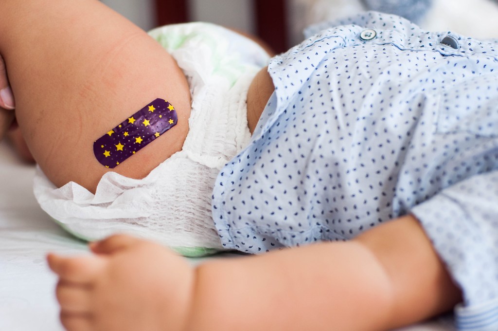 An infant lies on a table with a patterned Band-Aid on their thigh.