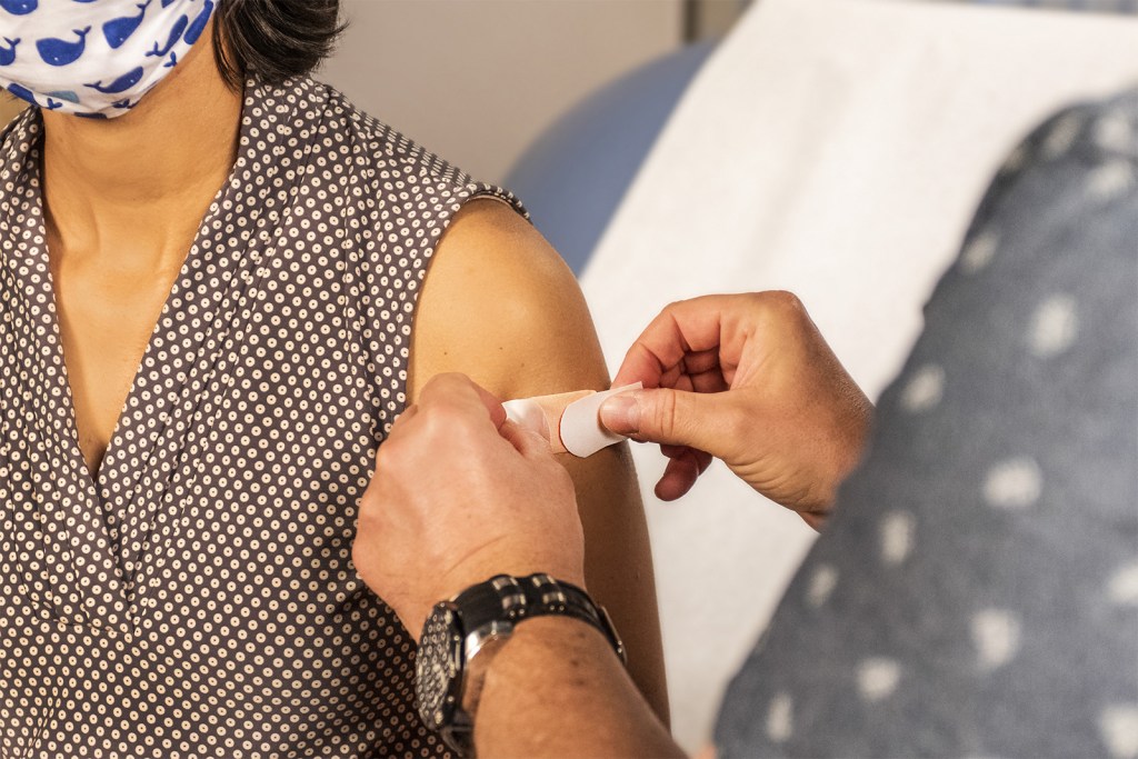 A woman wearing a cloth mask receives a bandage on her shoulder from a health care professional. 